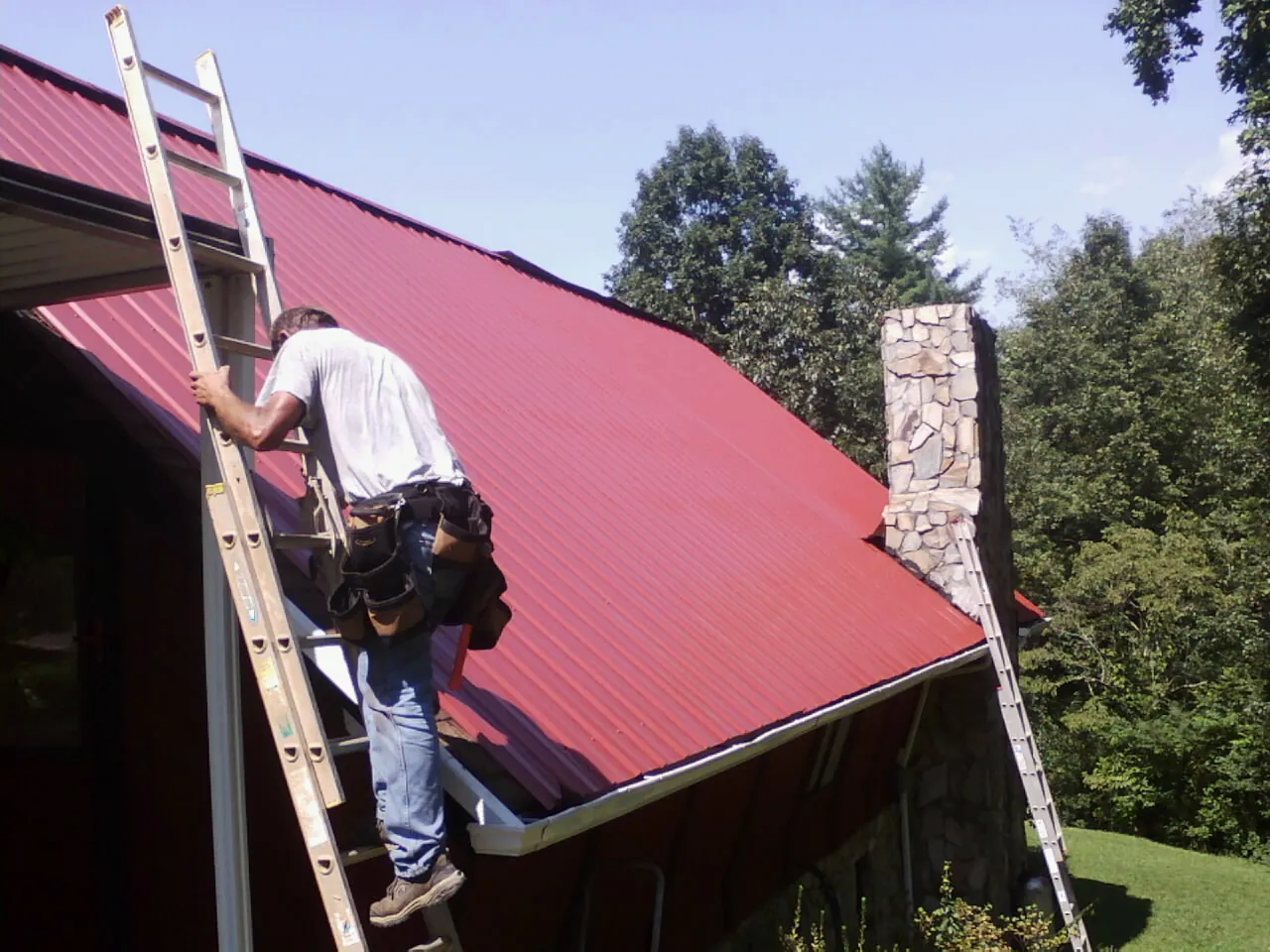 Roofer on a ladder beside a red metal roof and stone chimney — Soffit & Fascia Repair by Roofing Brothers in Talking Rock
